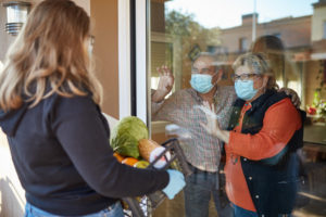Senior couple on their 70s wearing a protective face mask standing by the window indoors and watching their granddaughter delivering a basket with groceries in times of COVID-19, she is wearing mask and gloves.